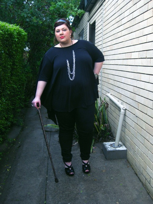 Outfit photo of me wearing a black drapey top with black skinny jeans and big black high heels with a platform. I'm leaning on my leopard print walking stick.