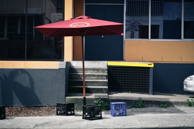 Photo of a cafe style umbrella on a footpath with  three milkcrate seats underneath it.