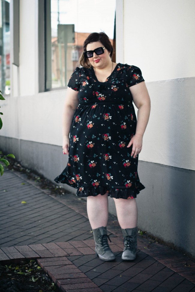 Outfit photo of me wearing a black babydoll style dress with short puffed sleeves in a small floral patterned fabric. I'm wearing it with grey boots and dark sunglasses.