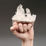 A photo of a fist clenched around a ring that surrounds all four knuckles with a huge lump of spiky crystals mounted on it.