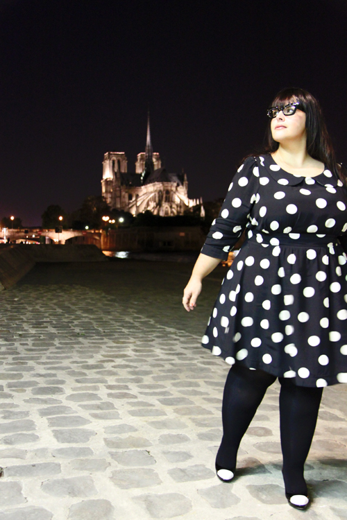 Photo of a fat young pale skinned woman with long dark hair and dark rimmed glasses wears the polka dot dress in front of what looks like a fancy building!