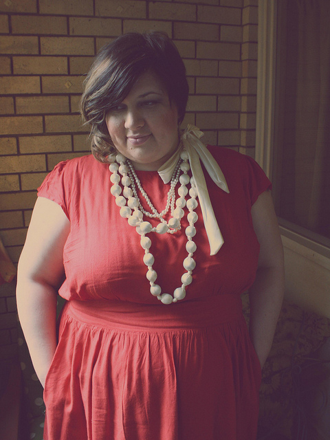 A portrait of me wearing a red dress and multi strands of pearl necklaces, one with a big ivory bow at the side of my neck. The colours in the photograph are faded and a bit dreamy.