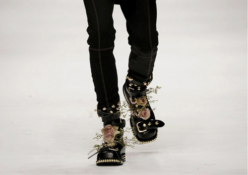 Photo of two black skinny jean clad legs walking down a white catwalk wearing big black boots with straps and pale pink roses with baby's breath tucked into the front of them.