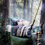 A photo of a bed with a handmade quilt and lots of cushions that is suspended by rope from trees in the middle of a forest.