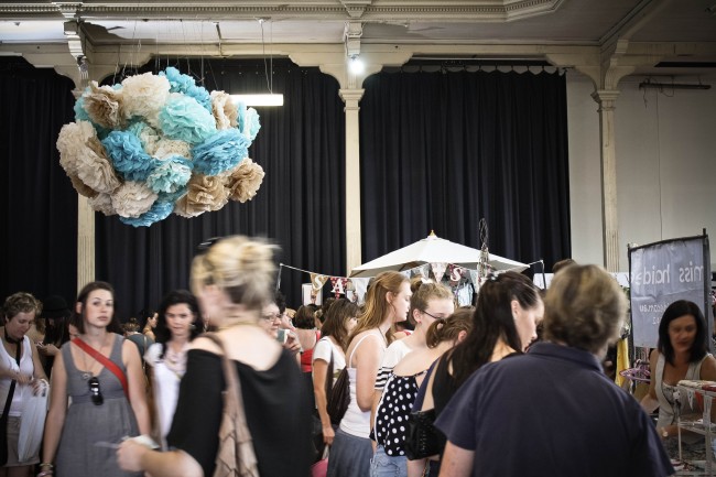 Photo of a large hall with lots of people around looking at cute craft shops. A cluster of huge pom poms hangs from the ceiling in the centre of the very large room.