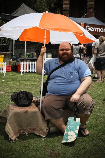 Photo of Nick, a pale skinned fat man with an amazing beard, sitting on a hession covered milk crate holding up a large orange and white umbrella.