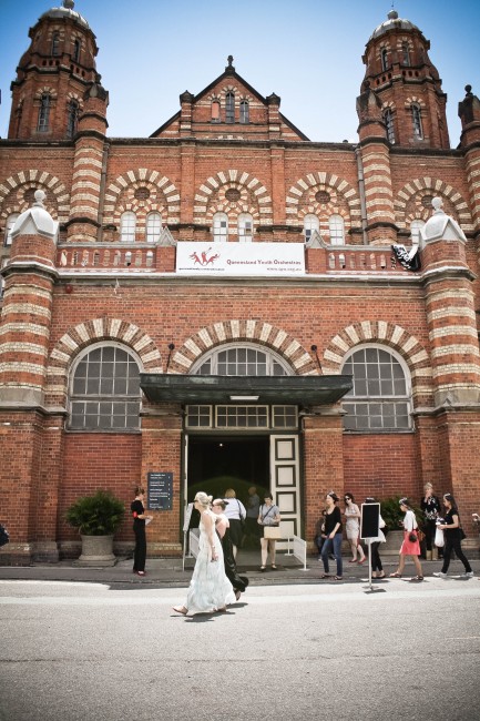 Photo of a large red brick hall entrance with decorative brickwork over arches and around columns. People are coming in and out of the door.