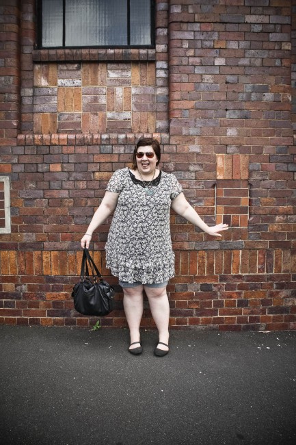 Photo of me against the side of a red brick exhibition building wearing a floral dress with denim cut off shorts. I'm making a pretty goofy face and pose.