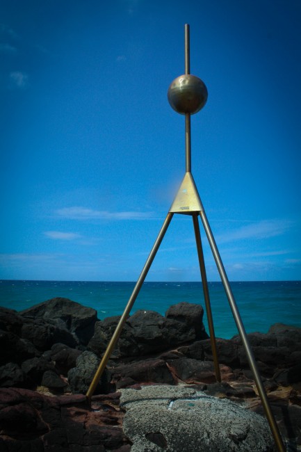 Photo of a tall metal structure which has a sphere sitting atop a tripod, it sits against a backdrop of rocks, the greeny blue sea and a bright blue sky with feint clouds.