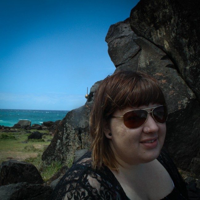 Photo of me (with a weird half grimace/ smile) sitting against rocks with the greeny blue sea and sky in the background.