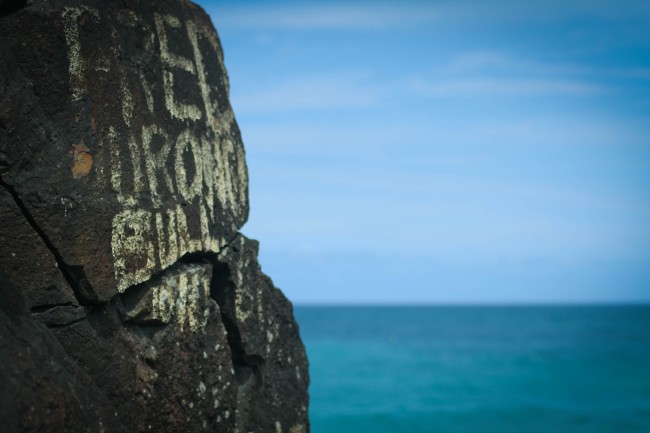 Photo of a huge rock with graffiti that is quite old and weather worn, the only legible word says "FRED". In the background is a blue green sea meeting the blue sky with feint clouds stretching across it.