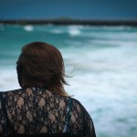 Photo of the back of me sitting on rocks facing the sea, I'm wearing a black lacey top and my hair is a little bit windswept.