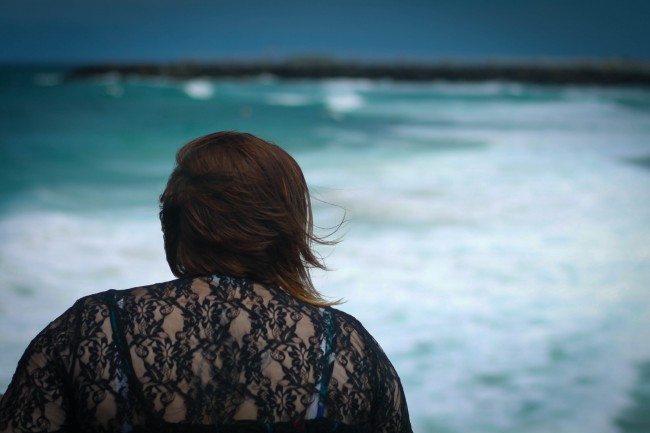 Photo of the back of me sitting on rocks facing the sea, I'm wearing a black lacey top and my hair is a little bit windswept.