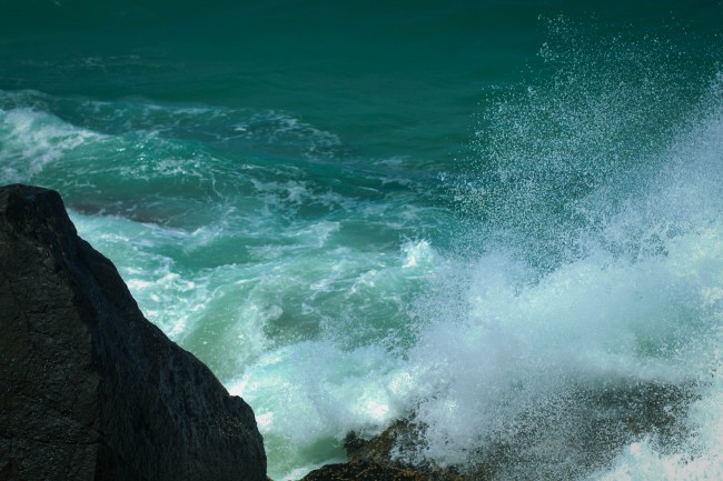 Photo of white foamy waves crashing against rocks, with the sea beyond a rich teal colour.