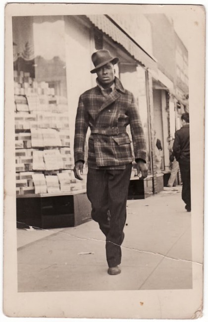 A sepia toned vintage photograph of a young slim dark skinned man wearing a checked jacket belted at the waist with a turned up collar, pin stripe pants and a jaunty hat. He is  strutting down the footpath with confidence but looking at the photographer questioningly.