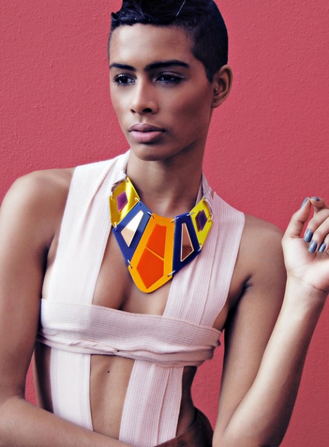 Photo of a young thin person with coffee coloured smooth skin wearing a bandage style top and a statement necklace made from coloured acrylic in geometric shapes that sit together like plates.