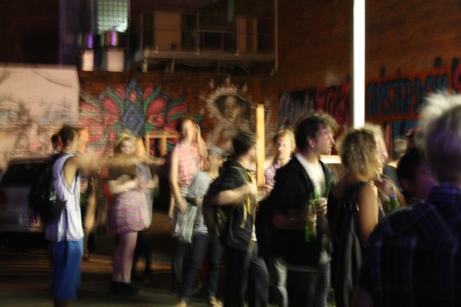 A blurry photo of people milling about in a carpark with heavily graffittied walls at night.