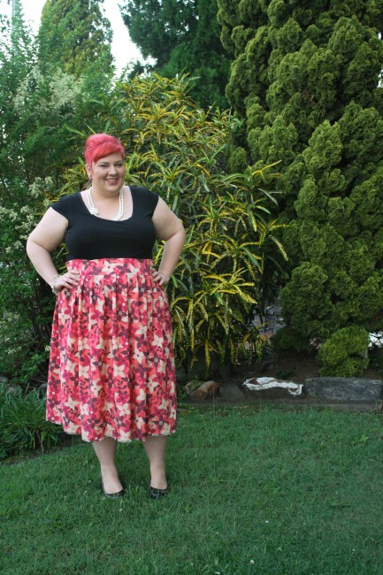 An outfit photo of me in the garden. My hair is pink and in a bouffant up-do; I wear a black top tucked into a high waisted and full skirted pink patterned skirt with a white necklace and black wedge court shoes.