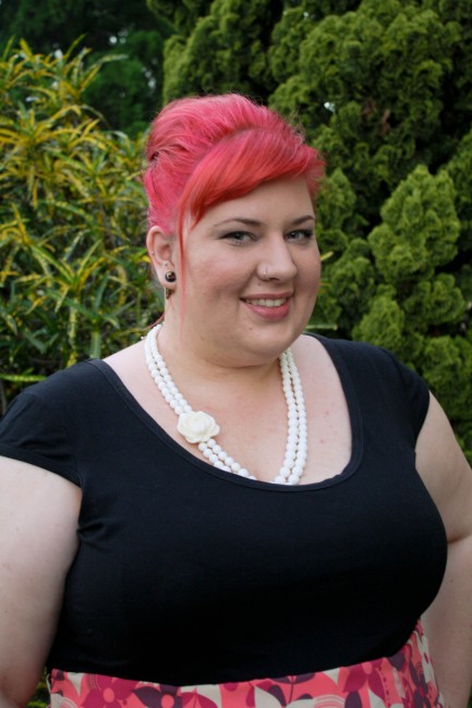 A head and shoulders photo of me  smiling with pink hair in a bouffant and a white bead necklace against a black short sleeved top.
