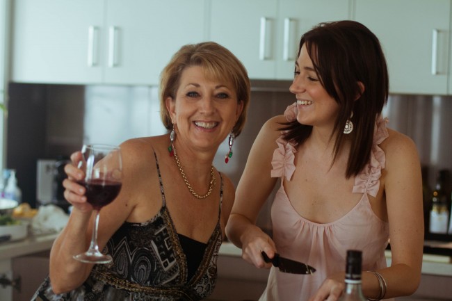 Photo of my mother, a middle aged lighted skinned woman with short light hair, and sister Amanda, a young slim dark haired woman, in the kitchen. Mum is drinking wine and smiling and amanda is holding a knife and looking at Mum and grinning.