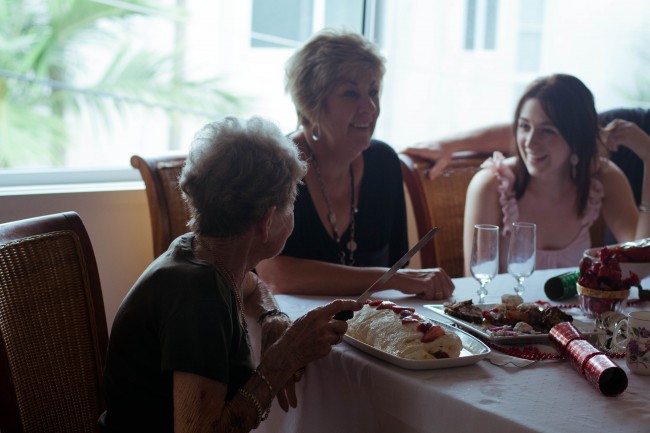 Photo of my Nana, an older pale skinned woman, and my Auntie Sharon, a middle aged pale skinned woman, and Amanda, young slim woman, at the table. Nana is holding a knife and getting ready to cut the pavlova that in front of her.