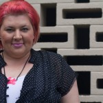 A head and shoulders photo of me, wearing a black shirt with polka dots over a white singlet with a pink fat necklace. I'm wearing awesome pale pink lipstick!