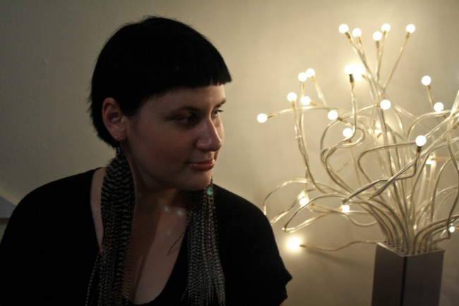 Photo of Sonya wearing lovely long feather earrings looking to the side looking pensive with a bunch of led lights behind her.