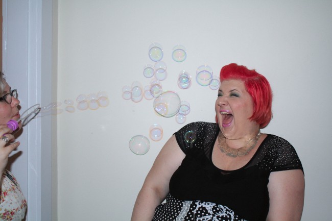 Photo of me making a kind of screachy happy face leaning back as Zoe blows bubbles at my head.