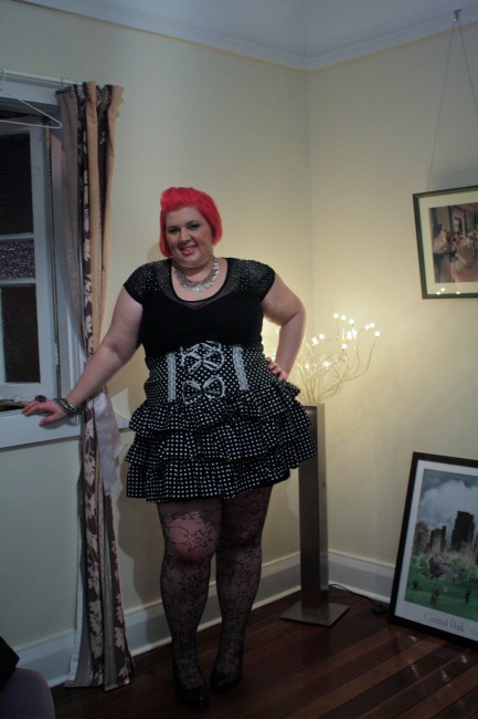 Outfit photo of me wearing a black top with a sheer yoke and shiny rhinestones on the shoulders, a highwaisted black and white polka dot ruffle skirt, lace fishnet tights and black shoes. 