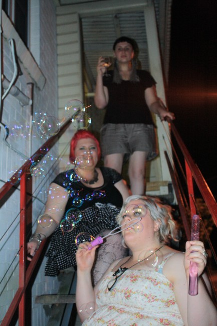 Photo of three women on an outside staircase at night: Zoe sitting in front blowing bubbles, me behind her smiling, and Sonya standing up at the back drinking wine.