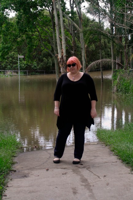 Photo of me standing in front of a flooded street. I'm wearing a black tunic with jeans and my hair is a faded pink; behind me are trees extending out of the water.