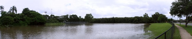 A panoramic photo of a park that has been filled with brown water. To the left are cricket cages, in the background are trees on the right side is a road and rail bridge.