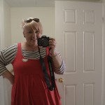 A photo of me looking into a bathroom mirror holding a DSLR. I'm wearing a black and white striped long tshirt under a pink pinafore with a white half doily brooch. My hair is now white-ish blond with an undercut and a long top and my sunglasses sit in my hair.