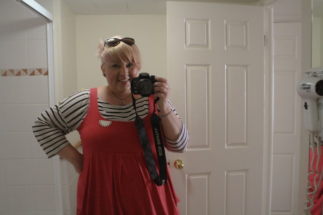 A photo of me looking into a bathroom mirror holding a DSLR. I'm wearing a black and white striped long tshirt under a pink pinafore with a white half doily brooch. My hair is now white-ish blond with an undercut and a long top and my sunglasses sit in my hair.