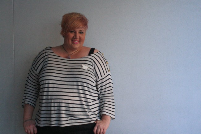 Photo of me (from the hips upward) standing against a white wall wearing a black and white striped tee with a half doily porcelain brooch.