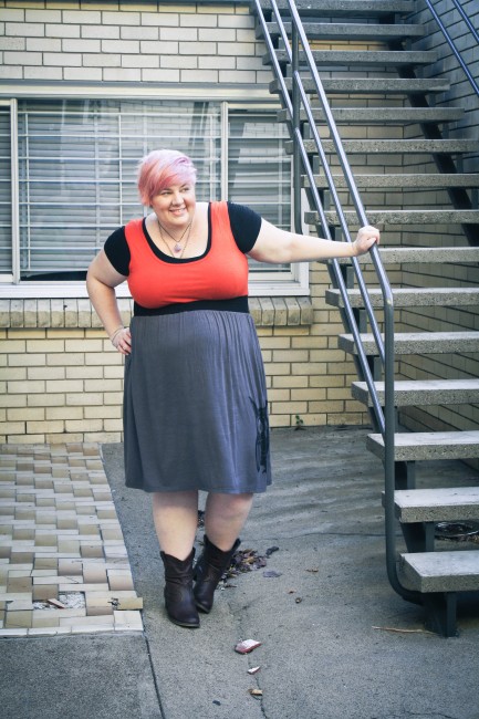 Outfit photo of me wearing the red and grey owl dress with black tshirt underneath and cowboy boots. I've got one hand on an external stairway rail and the other hand on my hip.