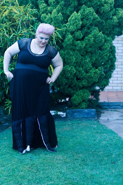 An outfit photo of me, fat and pale skinned, wearing my Ursula costume consisting of a black top with sheer sparkly shoulders and a long black maxi skirt with stuffed tentacles haphazardly attached to the bottom third of it. My short hair is sticking straight up with some white hairspray on the ends. 