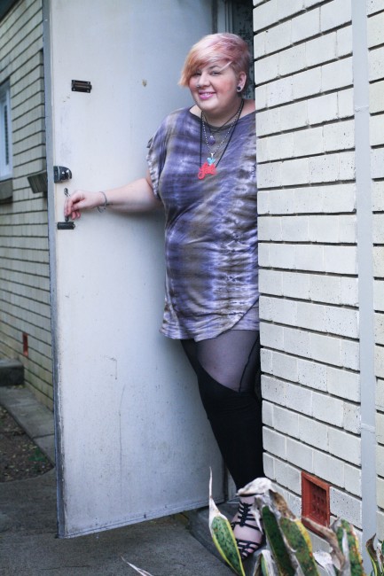 Outfit photo of me, a fat pale skinned lady, emerging from a door. I'm wearing a purple and grey tie dyed tshirt with black leggings with sheer cut outs as well as a pink fat necklace.