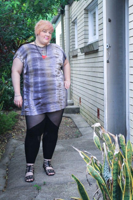 Outfit photo of me, a fat pale skinned lady, posing with one hand on my hip. I'm wearing a purple and grey tie dyed tshirt with black leggings with sheer cut outs as well as a pink fat necklace.