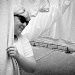 A black and white photo of me wearing sunglasses and peering around a towel on the line.