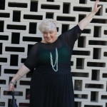 A photo of me, fat and pale skinned with short blonde hair, wearing a sheer black top with green singled under neath and large chain necklace. My arms are outstretched so you can see the baggy sleeve of the loose knit sheer top.