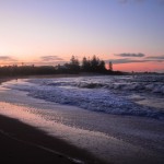 A photo of the waves rolling in at sunset. The beach curves around from the left foreground to middle background and the sky is a gradient of purple to pink.