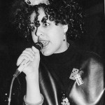 A black and white photo of Poly Styrene holding a microphone. She has dark short ringleted hair with a bow in it, braces on her teeth and is singing with big lovely wide-eyed intensity.