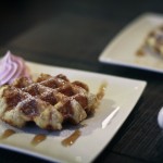 A photo of a square white plate with a delicious maple waffle drizzled in maple syrup and a little pink cream piped in the corner.