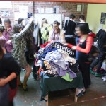 A photo of a room full of people moving about and holding up clothes that are piled on tables.