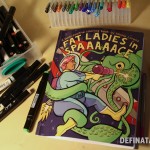 The front cover of Fat Ladies in Space, depicting a fat space lady battling with a space octopus as it tries to take hold of the space ship. Lots of markers and pens surround the colouring book.