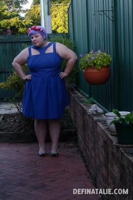 An outfit photo of me, fat and white skinned with short lilac hair and a pink flower crown, wearing the blue t-bar dress I made. I'm standing in our new yard!